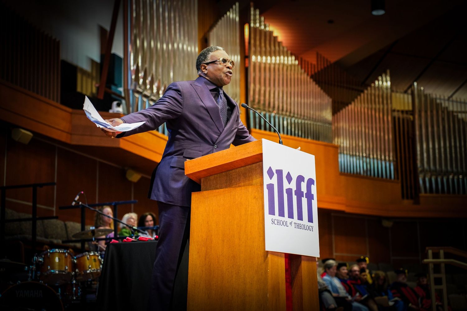 Passionate speaker addresses audience from ILIFF School of Theology podium in a grand auditorium.