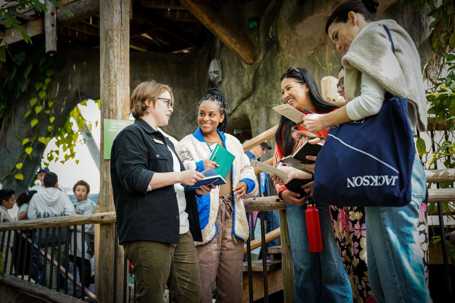 Group of four people smile and chat on wooden deck surrounded by greenery at outdoor event.