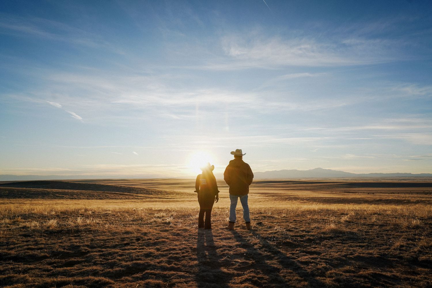Two people stand silhouetted against a stunning golden sunset over a vast open prairie landscape.