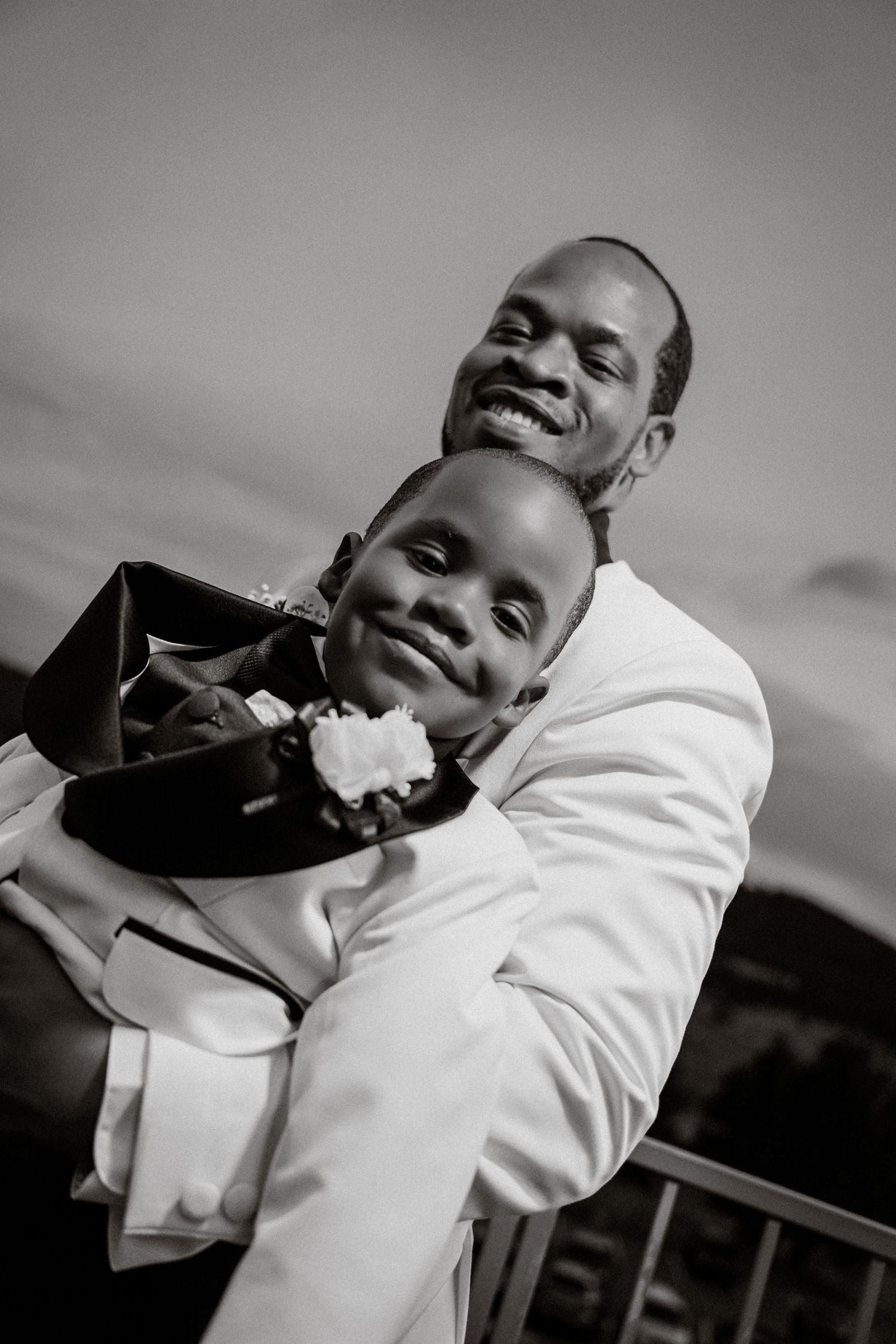 Black and white photo of a man in a white suit embracing a smiling boy in formal attire with a boutonniere.