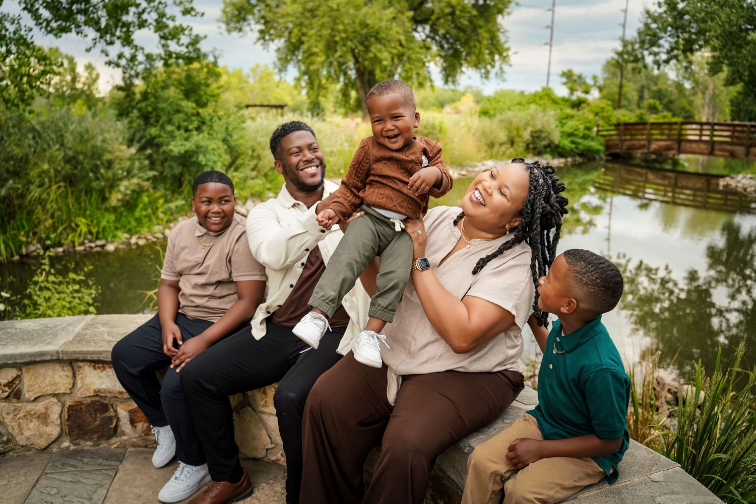 Happy Black family of five laughing together outdoors near a pond surrounded by lush green trees.