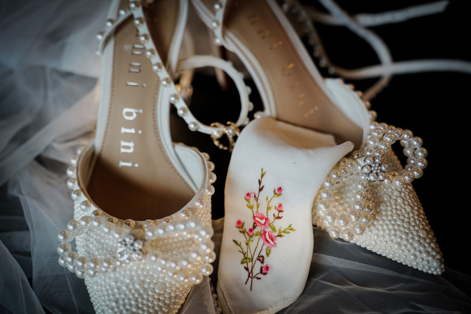 Elegant pearl-embellished wedding heels by Bini with floral embroidery detail, displayed on a white veil.