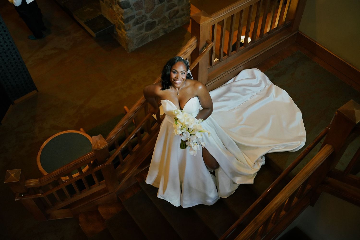 Bride in a stunning white ball gown poses on wooden staircase, holding a bouquet, captured from above.