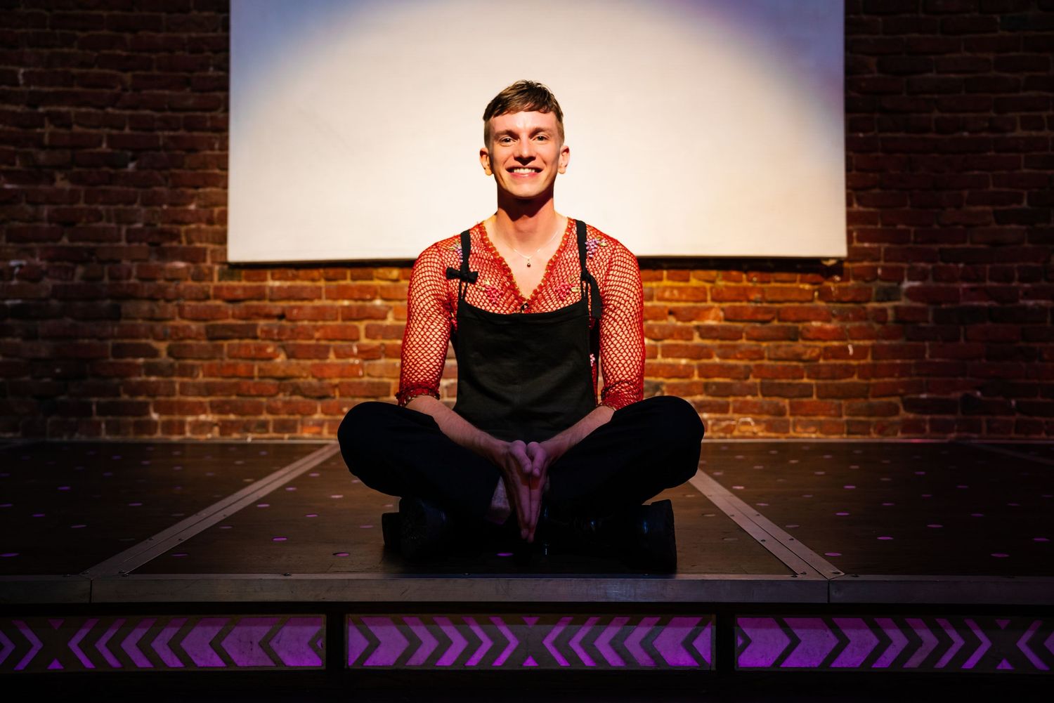 Smiling man in a red mesh shirt and black apron sitting cross-legged on a stage with purple arrow lights.
