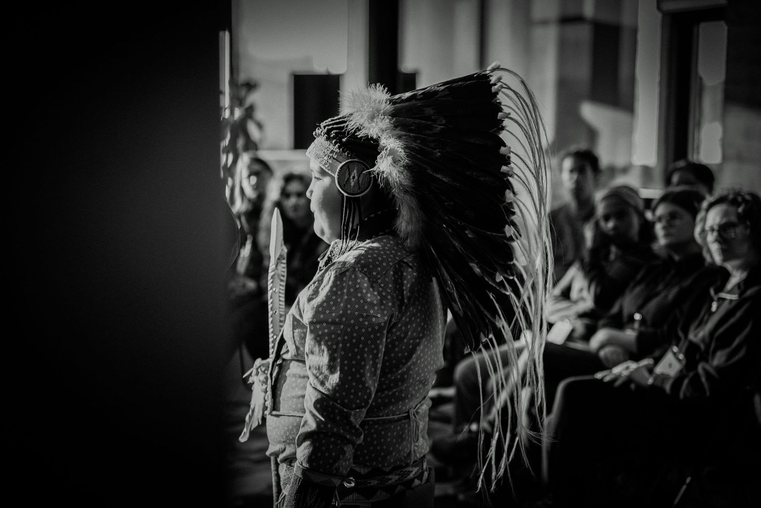 Black and white photo of person wearing Native American headdress presenting to seated audience indoors.