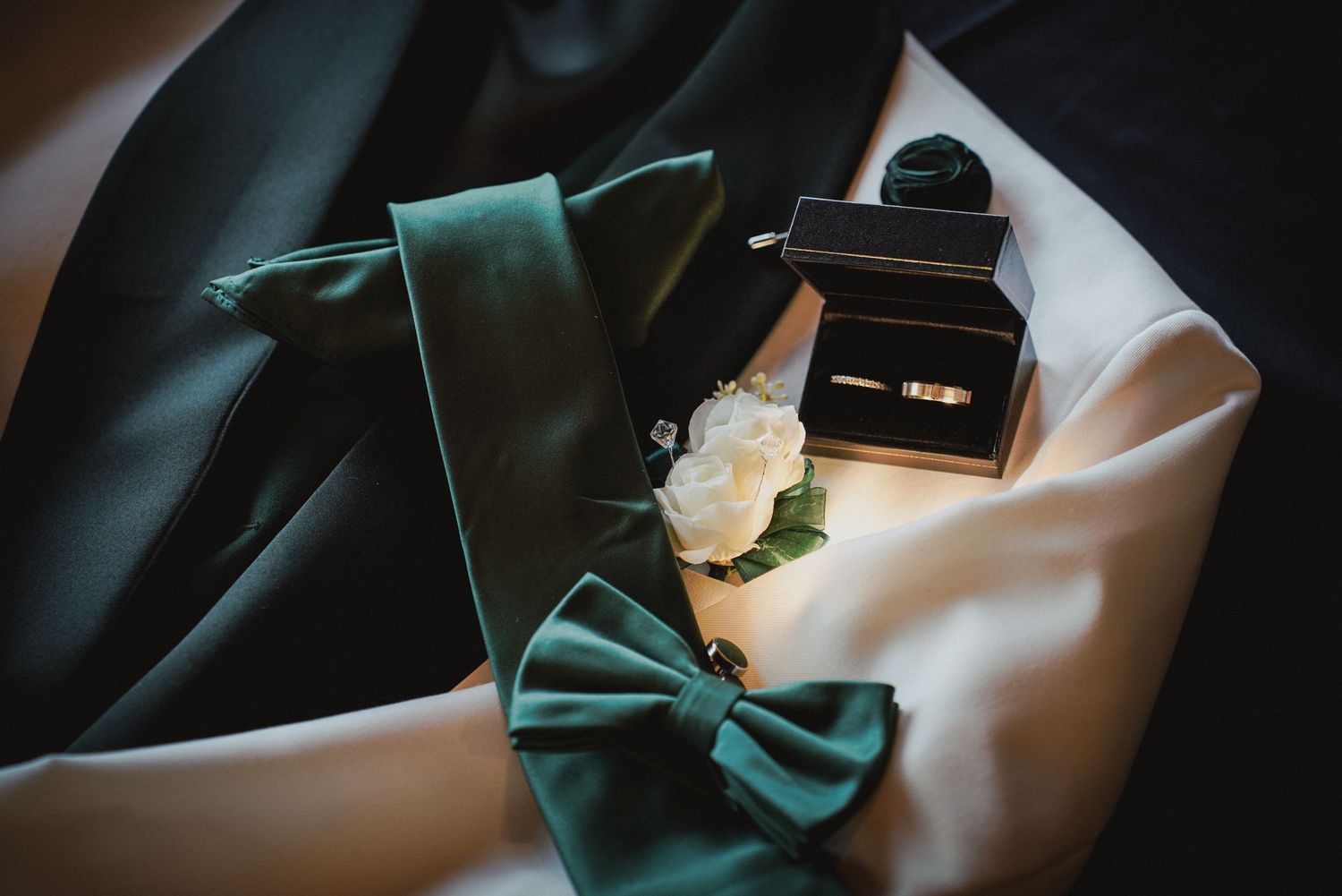Green bow tie and long tie with white rose boutonniere and gold wedding rings in box on white fabric.