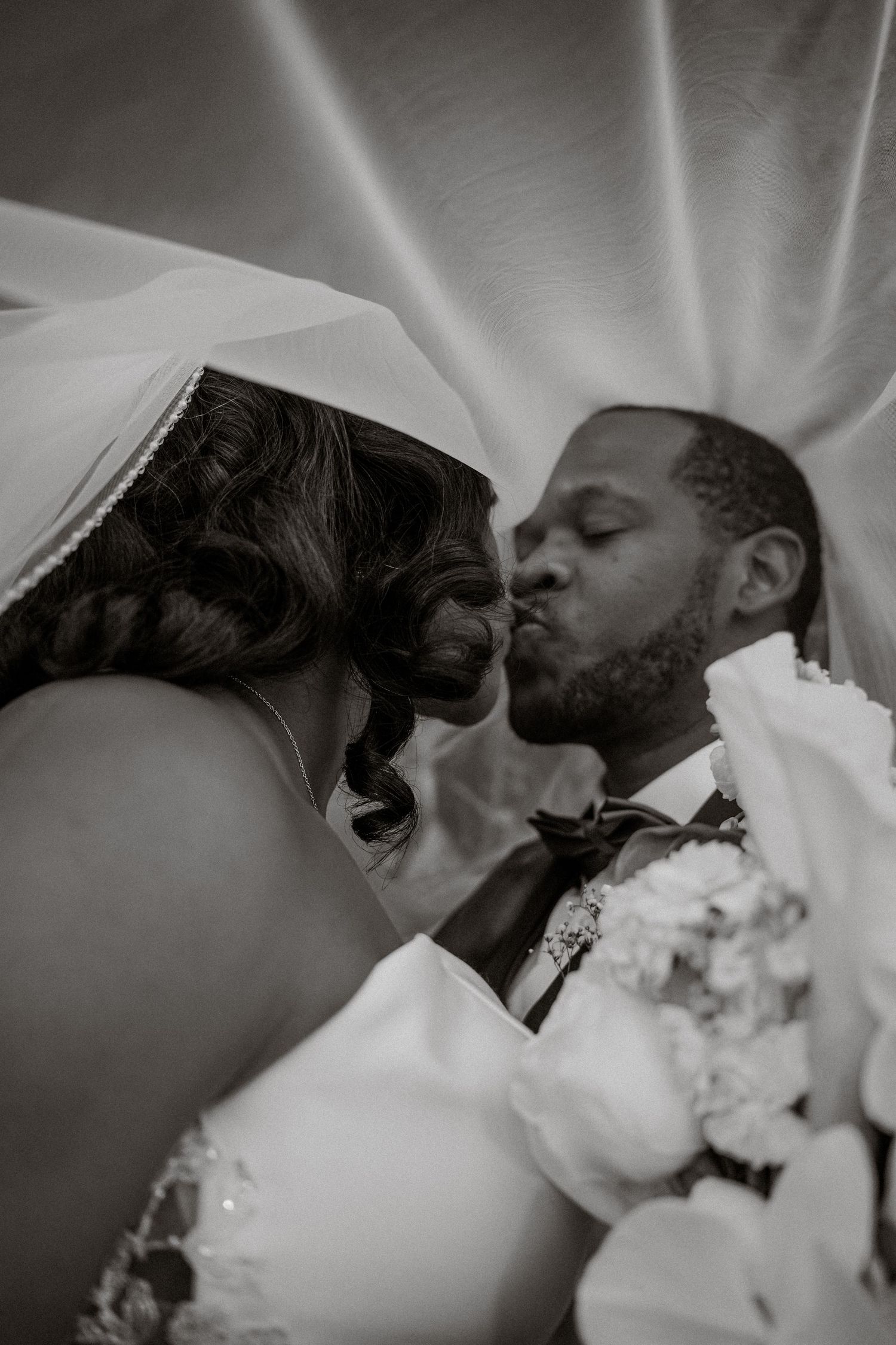 Black and white wedding photo of a bride and groom sharing a kiss under a veil.