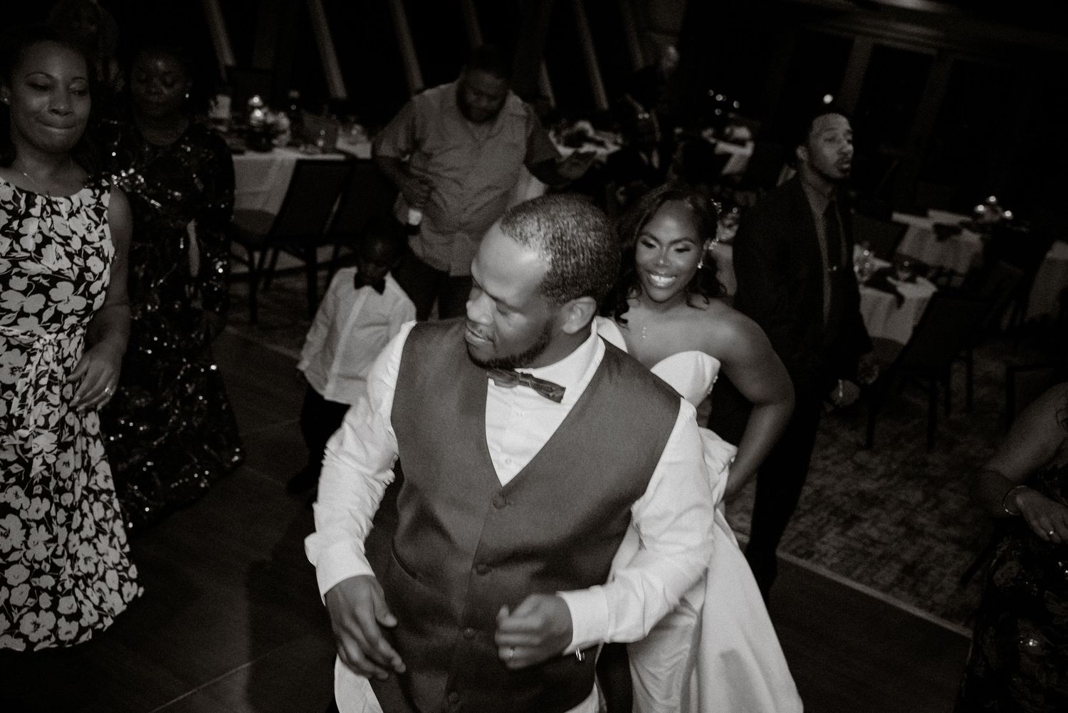 Black and white photo of a couple dancing at a wedding reception, groom in vest and bow tie, bride in white gown.