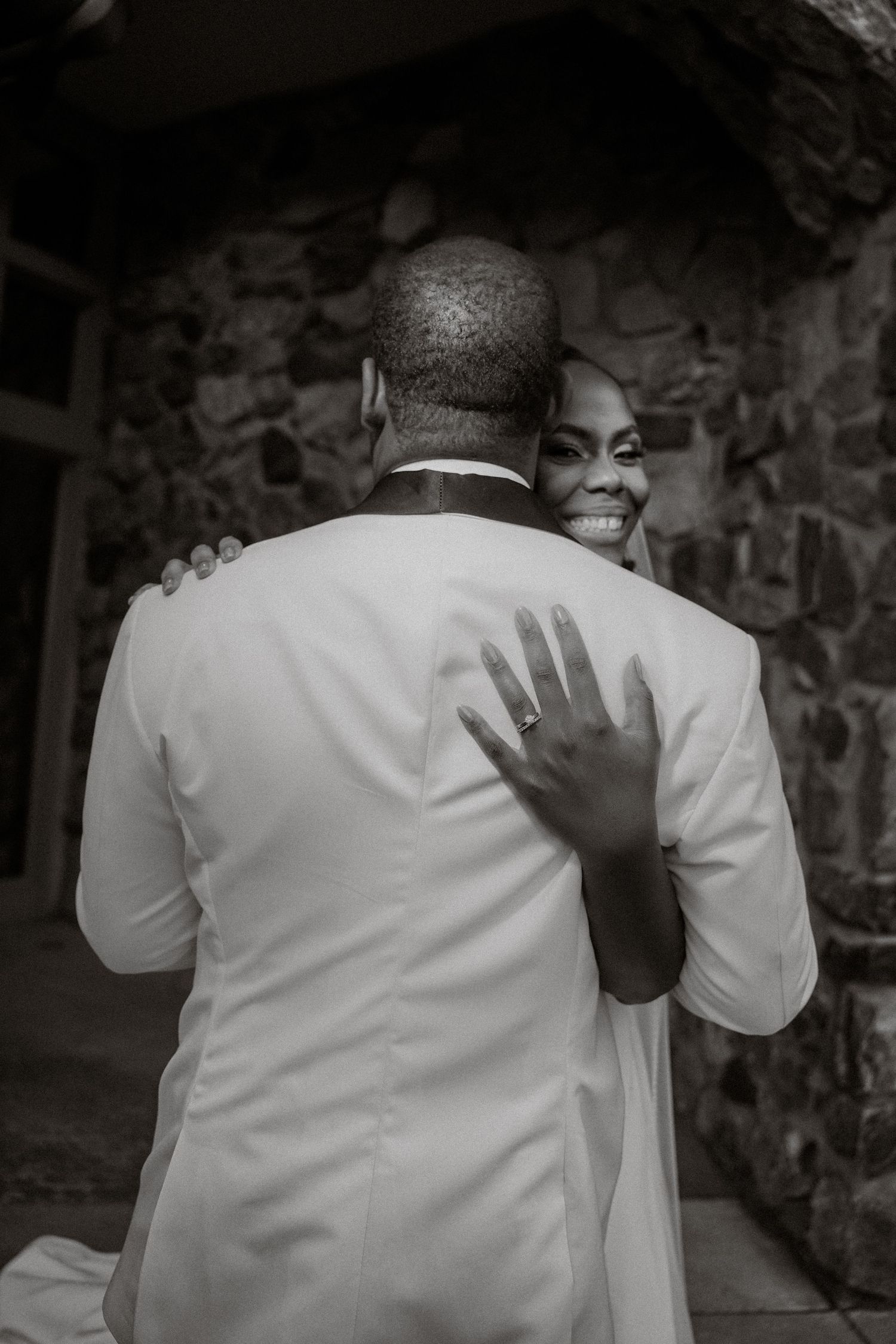 Black and white wedding photo of smiling bride showing ring while embracing groom in white suit near stone wall.