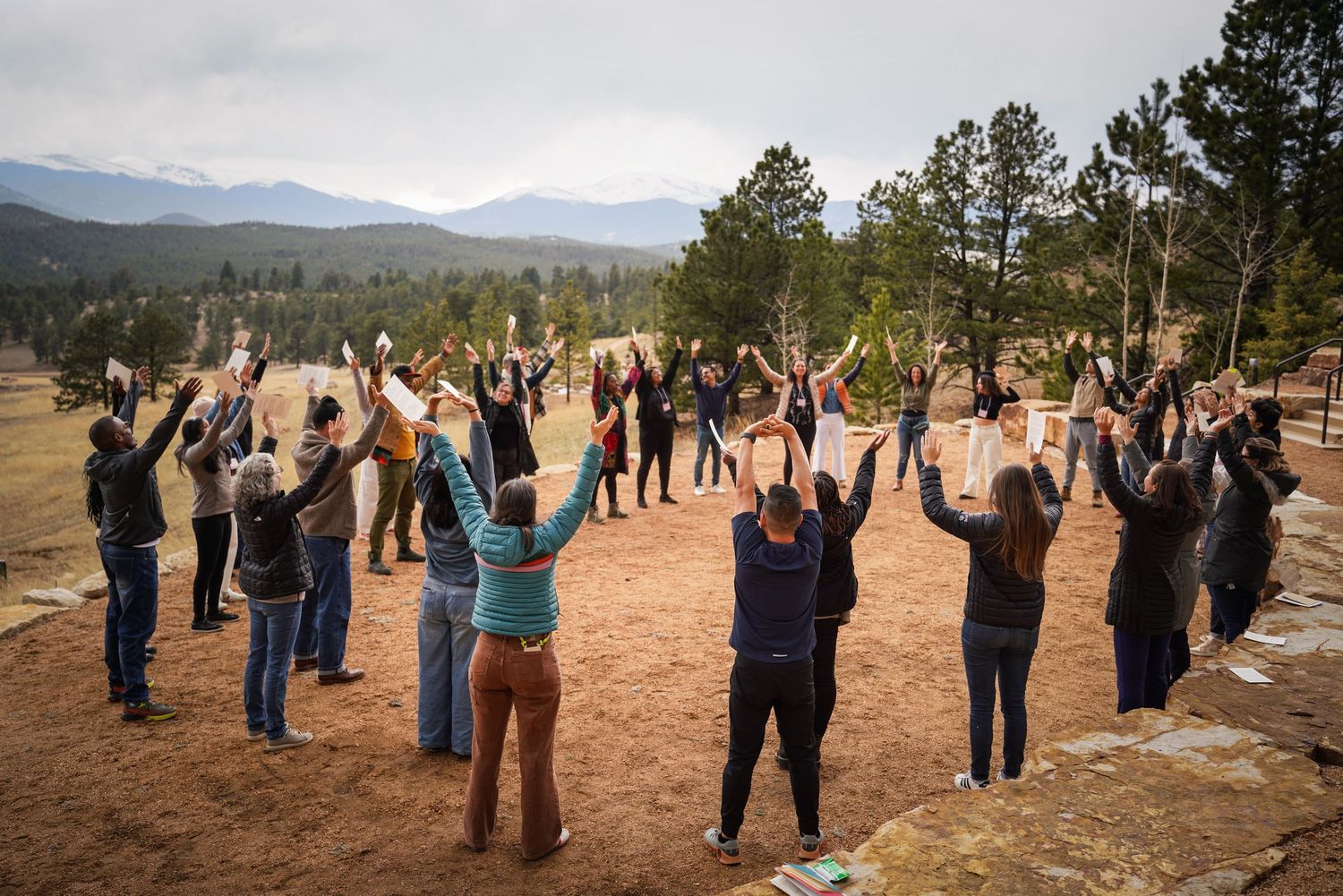 Large group of people standing in circle with arms raised during outdoor team building activity in mountains.