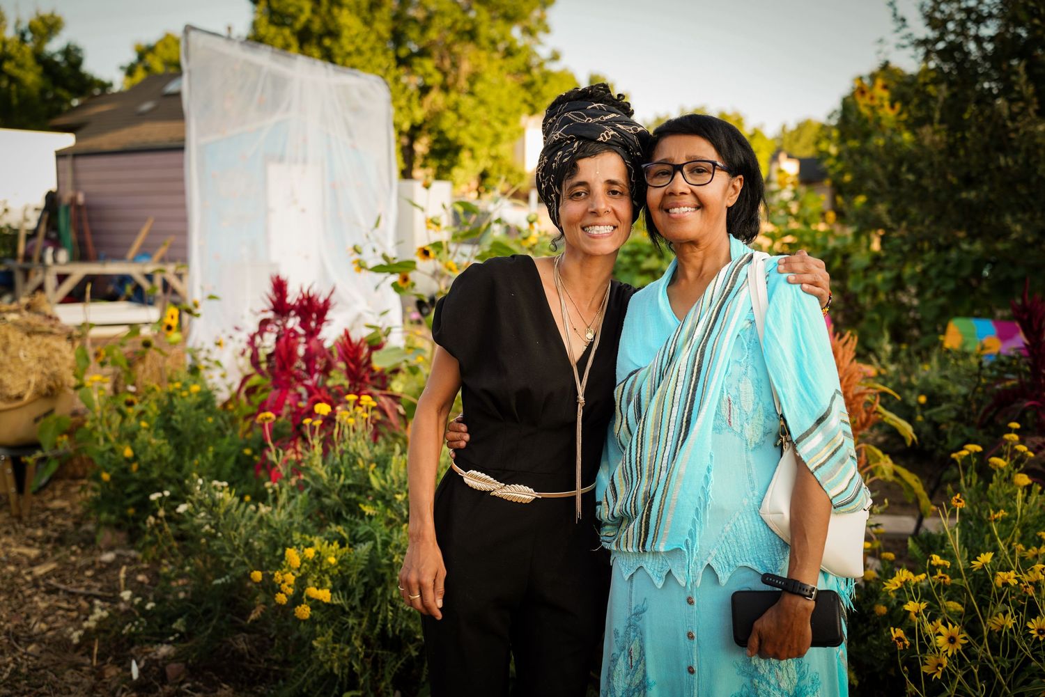 Two women smiling together in a vibrant community garden filled with colorful flowers at dusk.