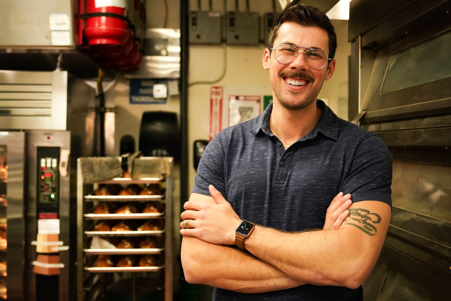 Smiling tattooed baker with crossed arms standing in a professional bakery kitchen.