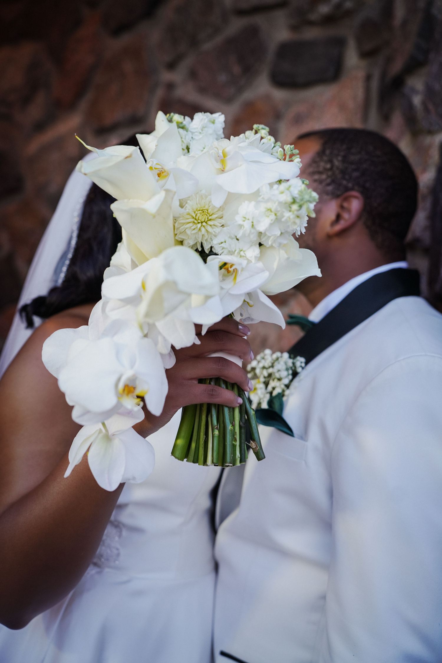 Bride and groom sharing a kiss behind a white orchid and calla lily wedding bouquet at their outdoor ceremony.