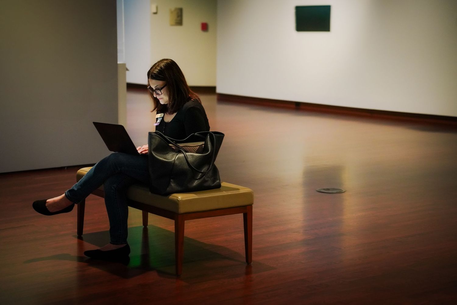Woman sits alone on bench using laptop in quiet museum gallery with artwork on walls.