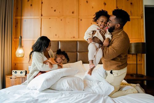 Happy family playing together on white bed in warm hotel room.