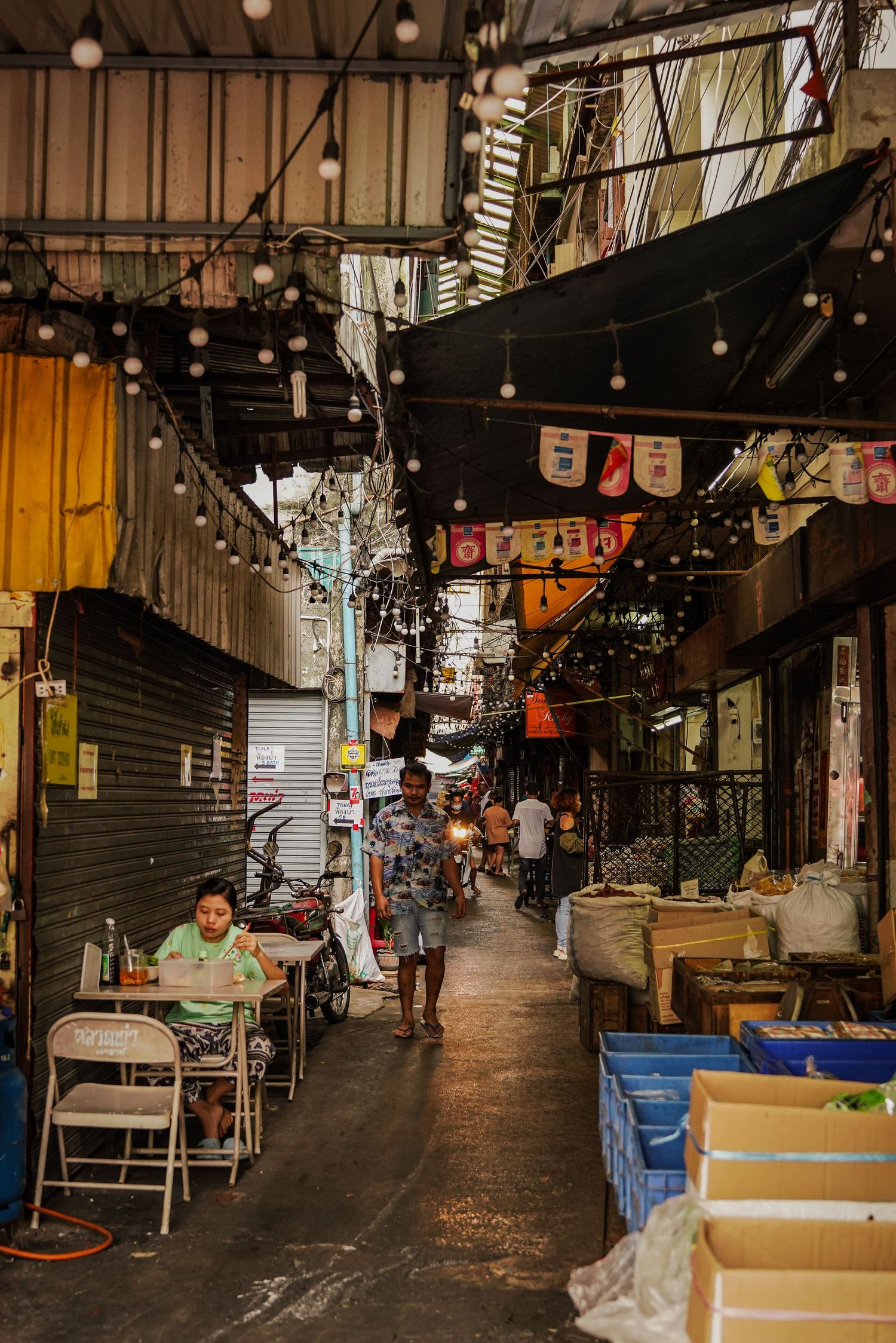 Narrow bustling market alley with string lights, shops, and vendors in urban Asia.
