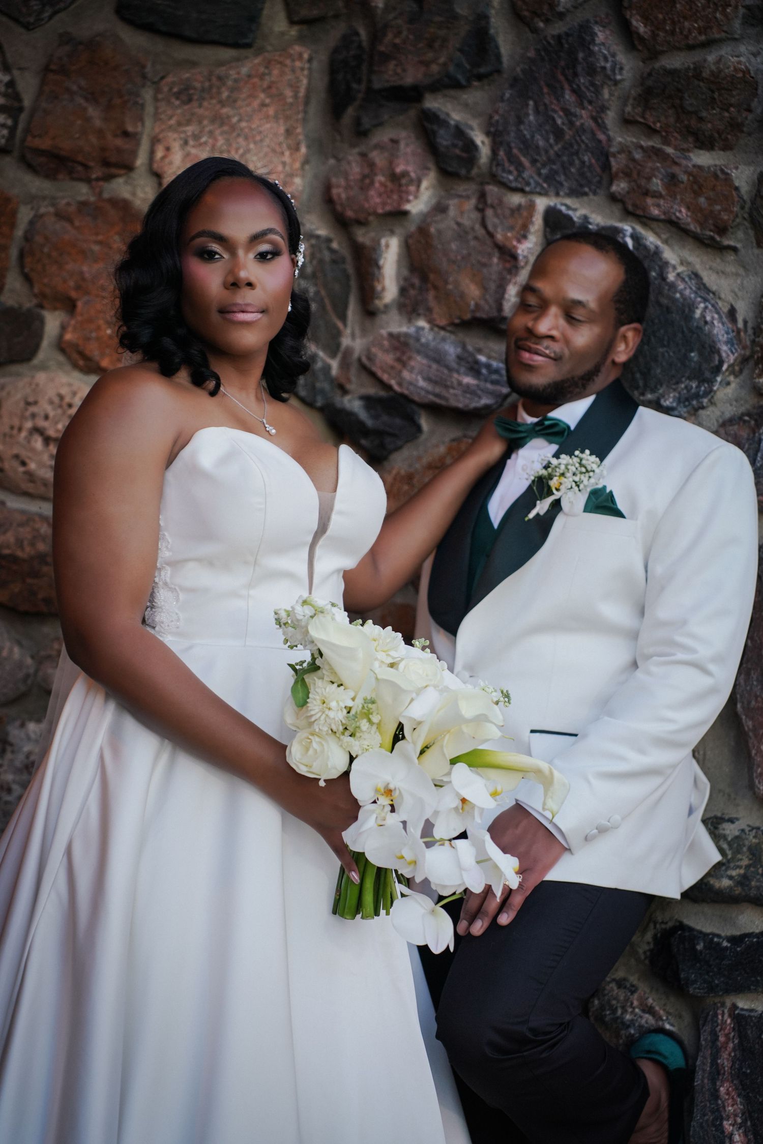Bride in white strapless gown holds white floral bouquet beside groom in white tuxedo jacket against stone wall.
