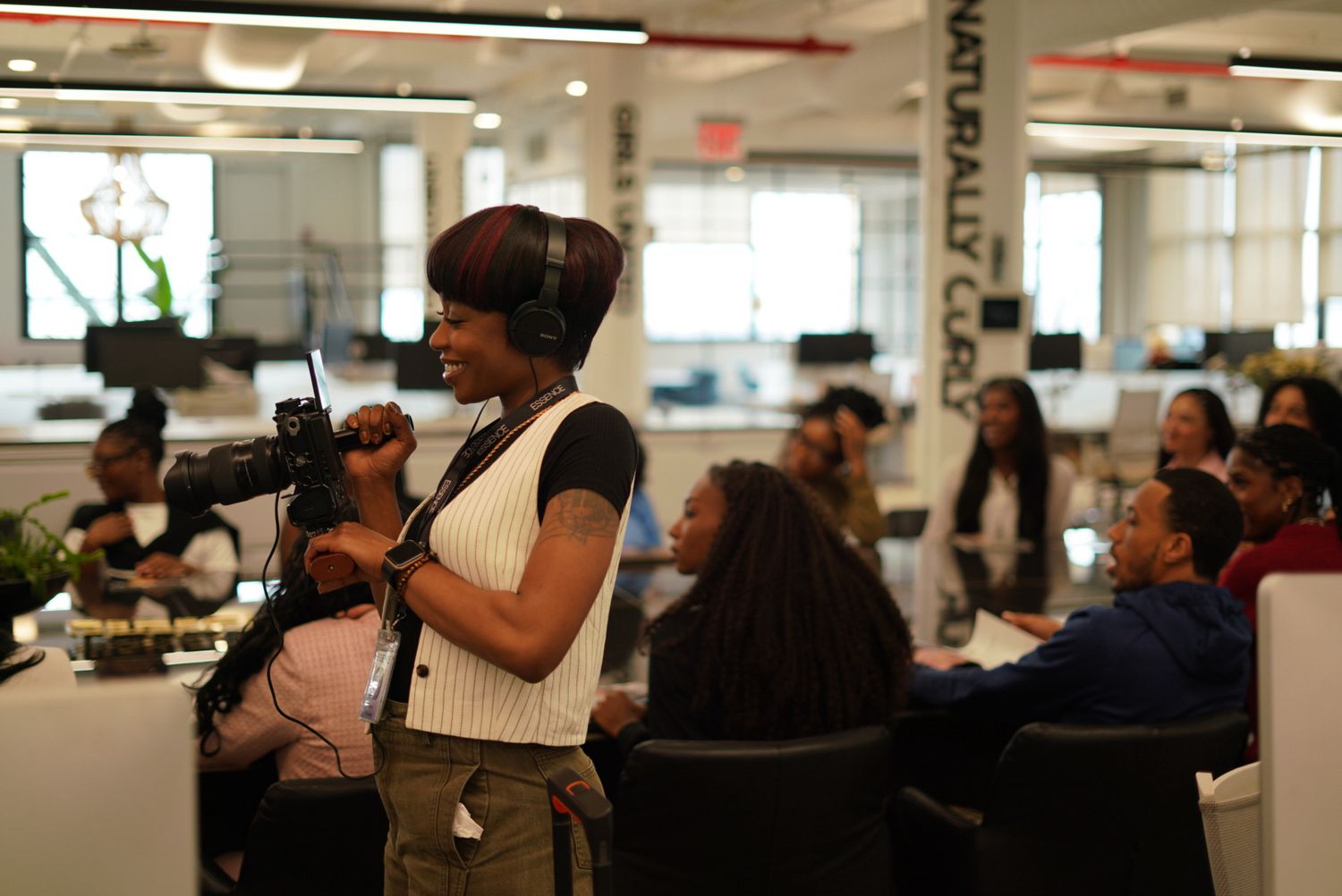 Woman with headphones holds camera in busy office, people with natural hair sit at tables in background.