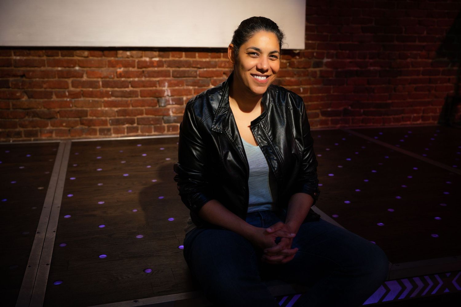 Smiling woman in a black leather jacket sitting cross-legged on a stage with purple lights and brick wall background.