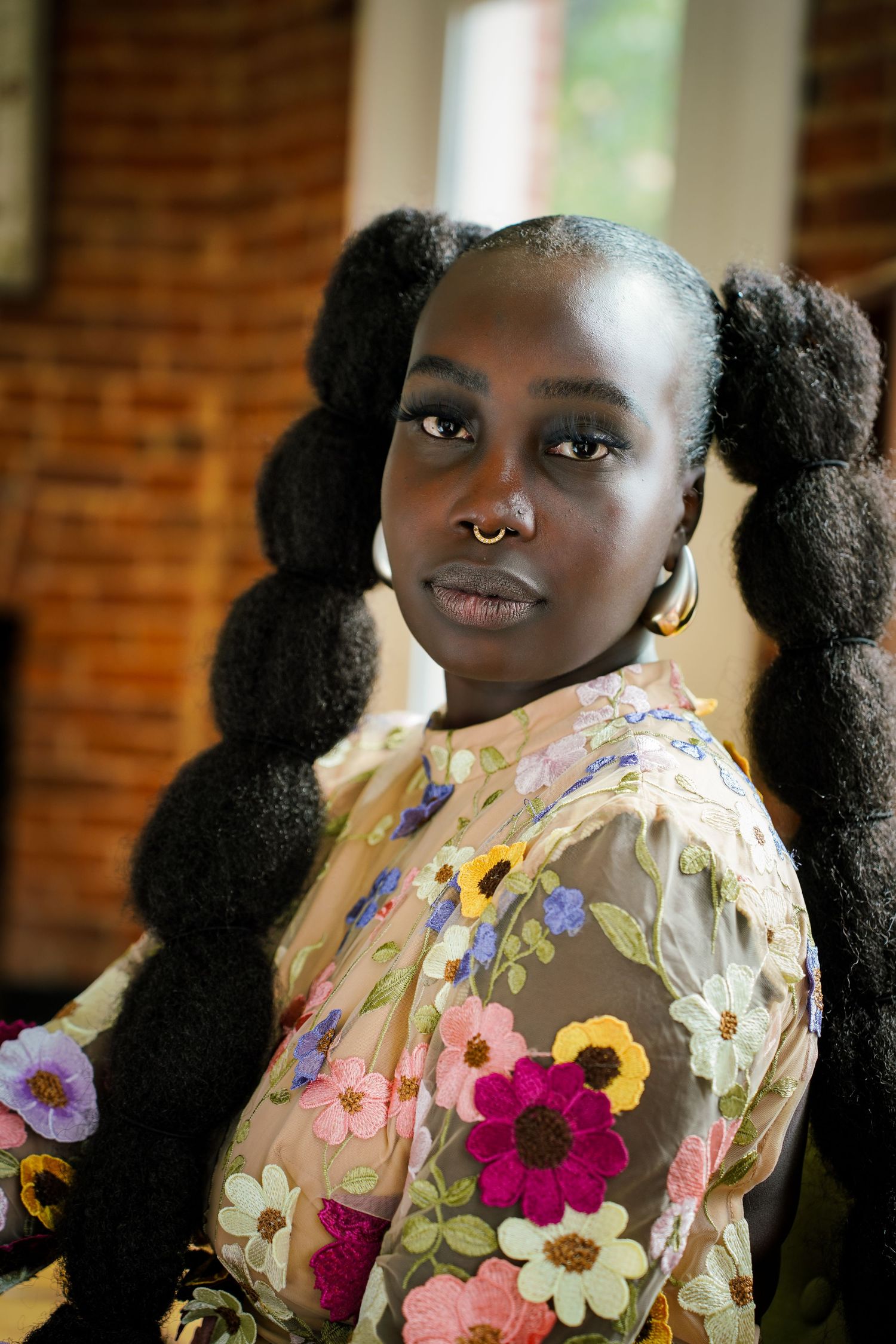Portrait of woman with large puff pigtails wearing floral embroidered sheer top near brick wall.
