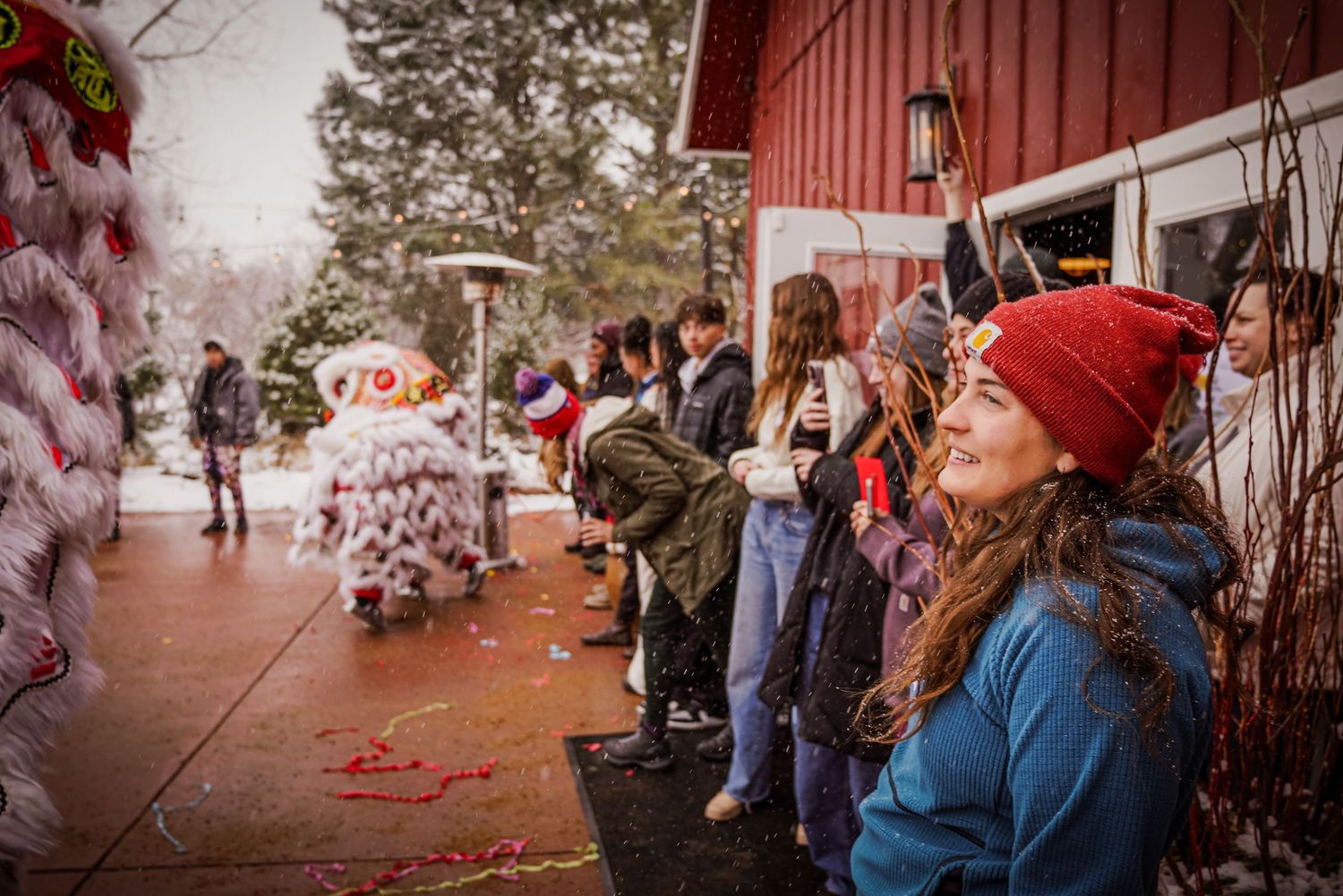 Smiling woman watches a traditional Chinese lion dance performance outside in snowy winter weather.