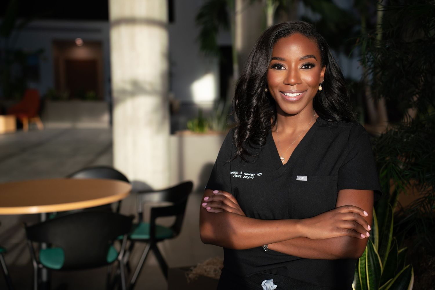 Confident woman in black medical scrubs stands with arms crossed outdoors at night near patio.