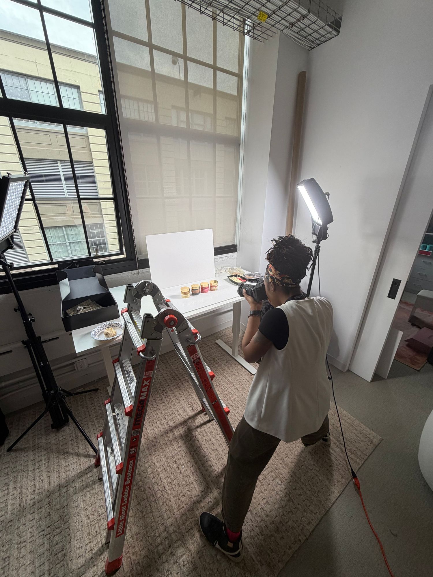 Photographer on ladder shooting product photography with LED light panel near large windows in modern studio space.