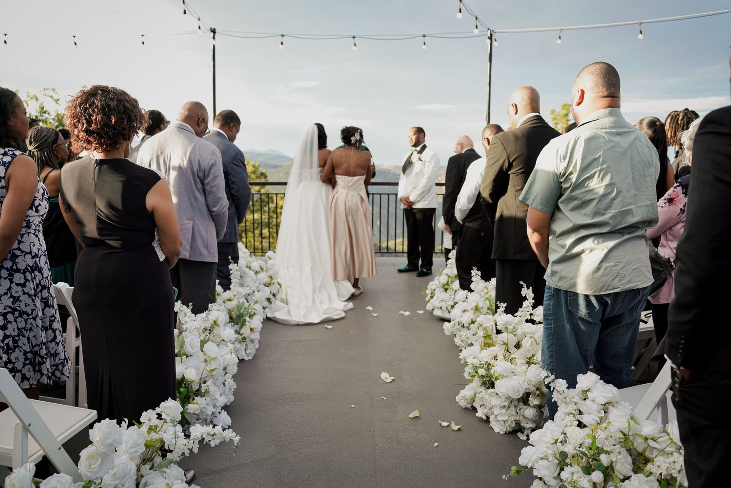Outdoor wedding ceremony on a rooftop terrace with white floral aisle arrangements and guests standing for the bride.