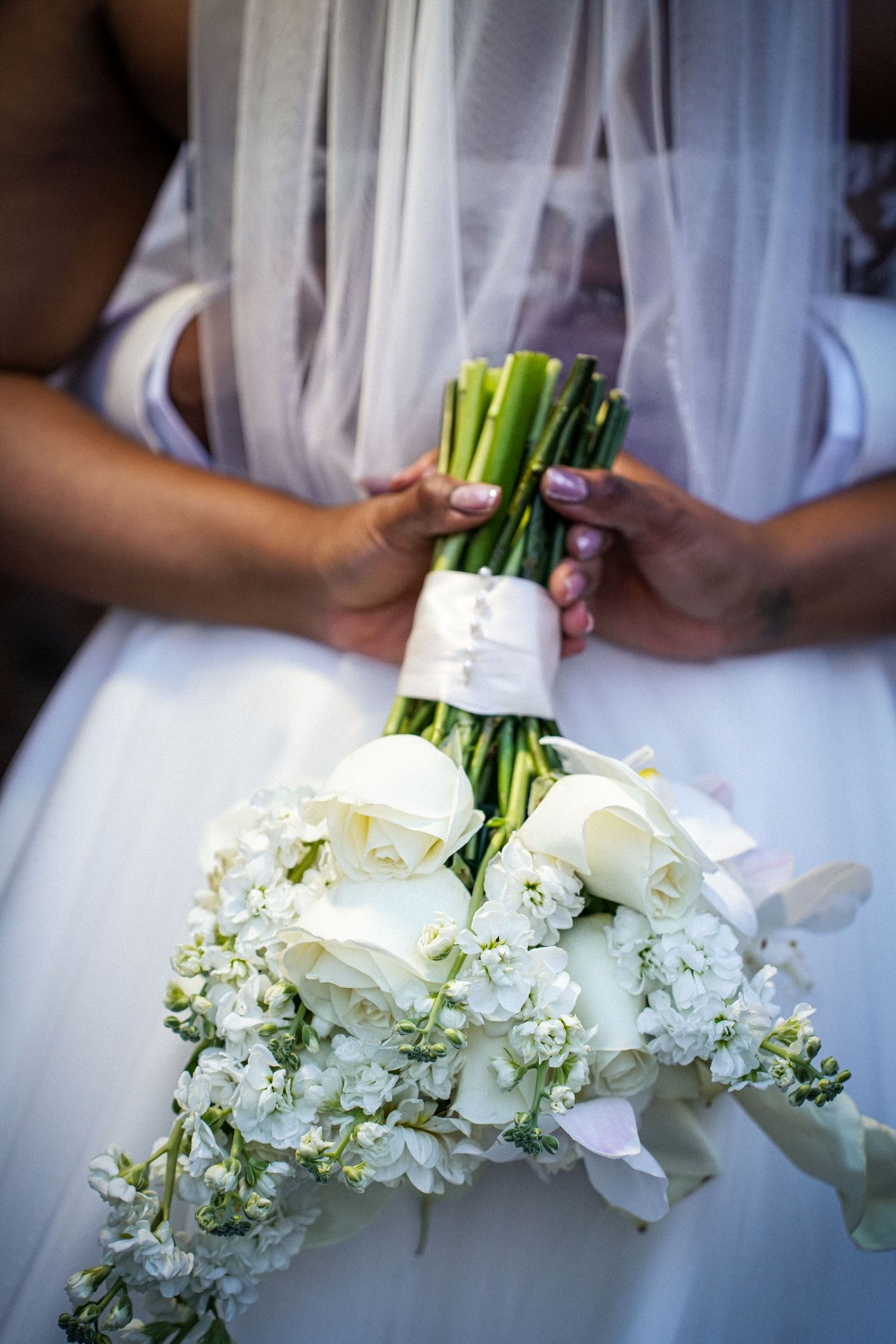 Bride in white wedding dress holding elegant bouquet of white roses and flowers wrapped with ribbon.