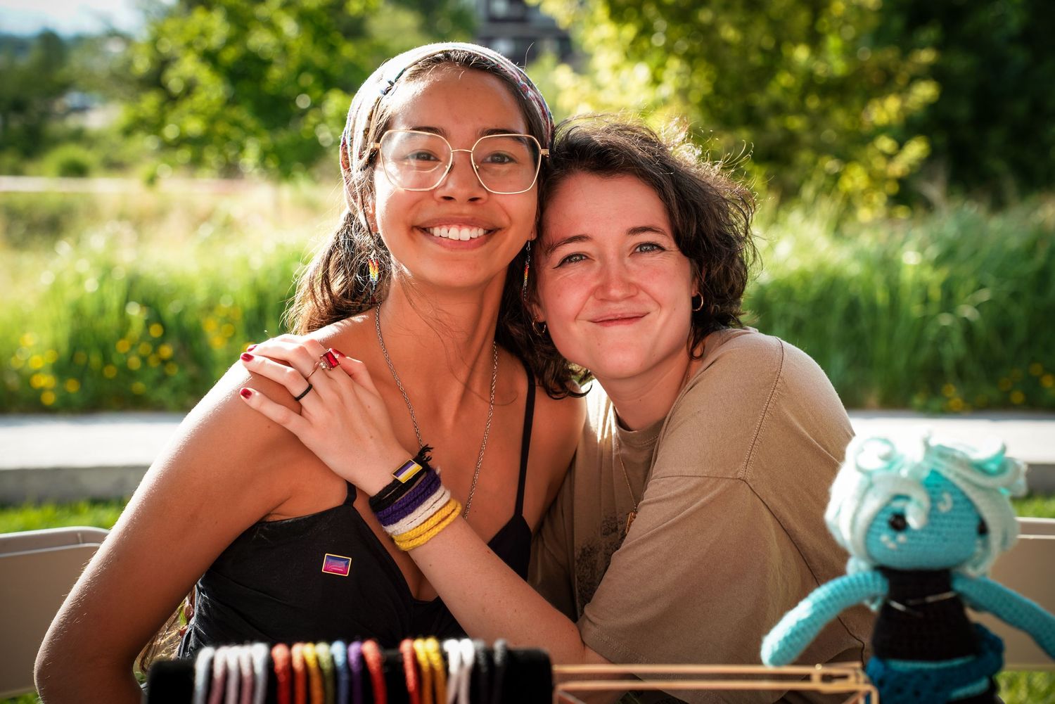 Two smiling young women embrace outdoors at a sunny Pride event with a teal dragon plush nearby.