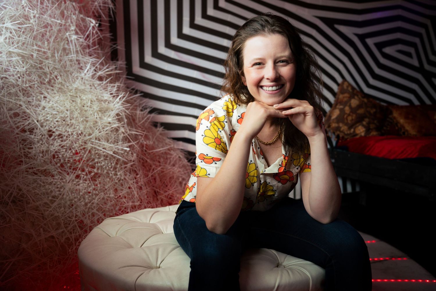 Smiling young woman with curly hair wearing a floral shirt, sitting on a tufted ottoman in a colorful striped room.