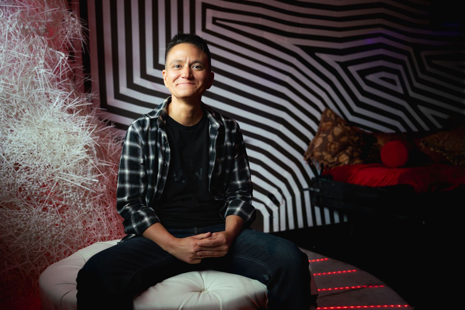Person in a plaid shirt smiling while seated on a white ottoman in a black and white striped room.