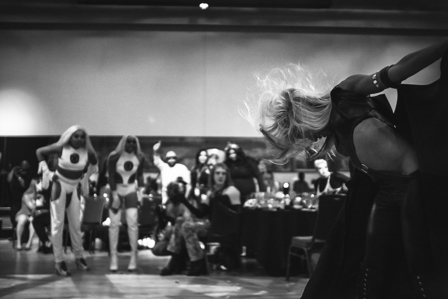 Black and white photo of performer tossing hair on stage before a seated audience.
