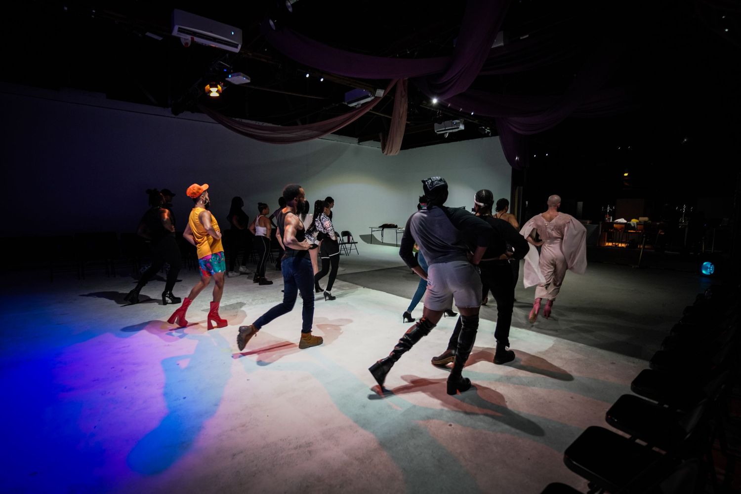 Diverse group of performers dance on colorful lit stage during theatrical performance in dark venue.