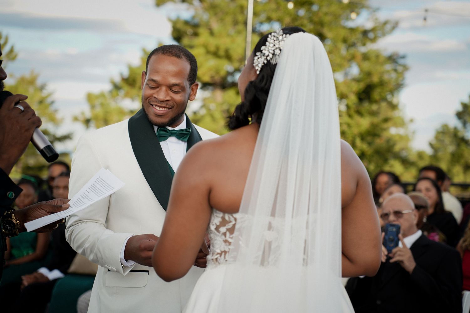 Groom in white tuxedo reading wedding vows to bride in lace gown and veil at outdoor ceremony.