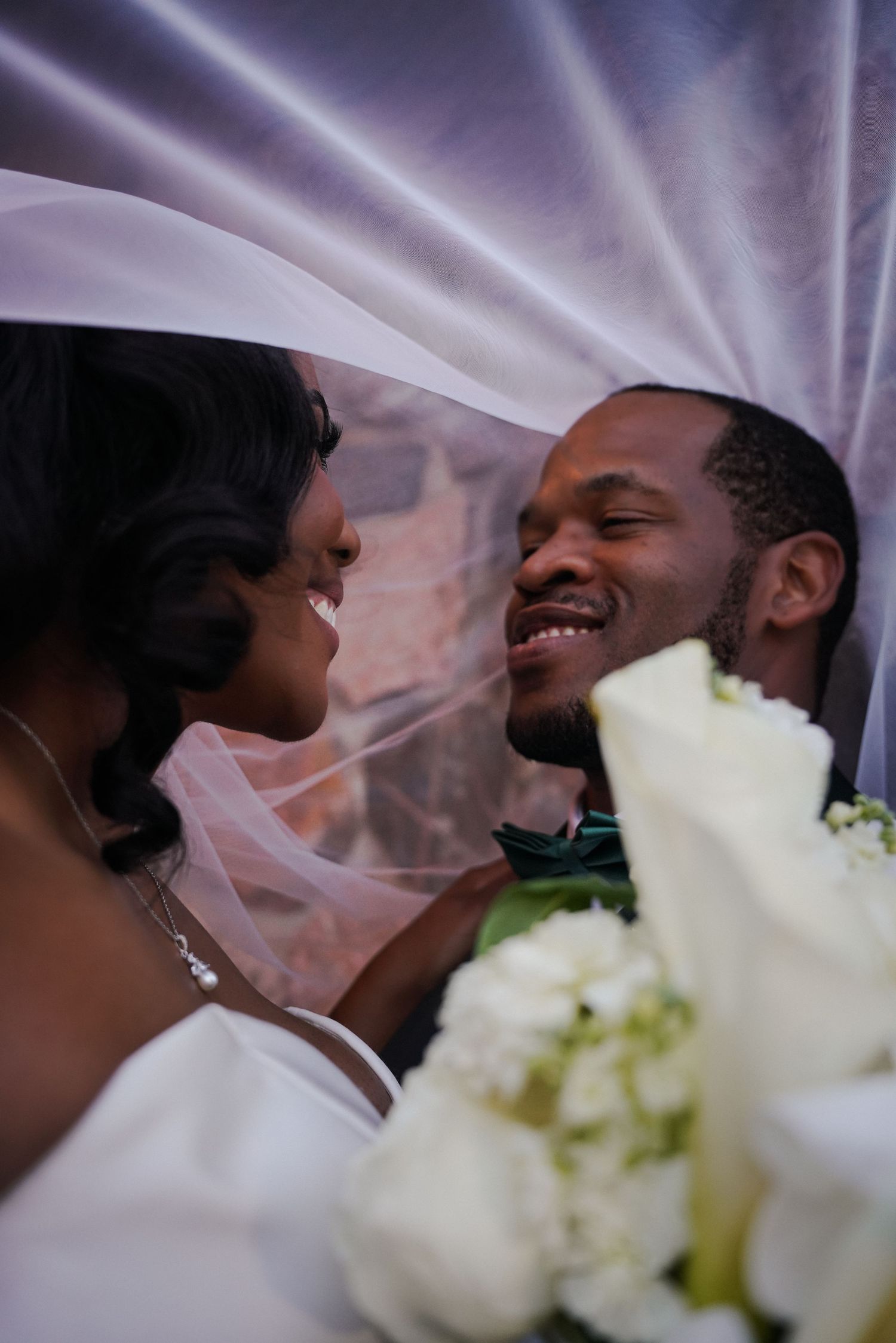 A joyful bride and groom share an intimate moment under a wedding veil, with white roses in the foreground.
