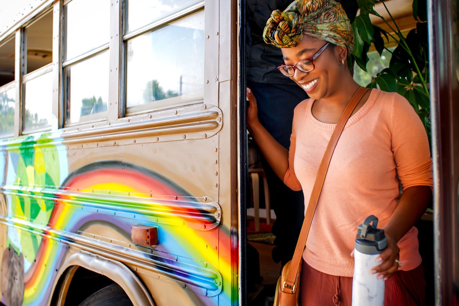 A smiling woman holding a water bottle standing beside a colorful rainbow-painted school bus.