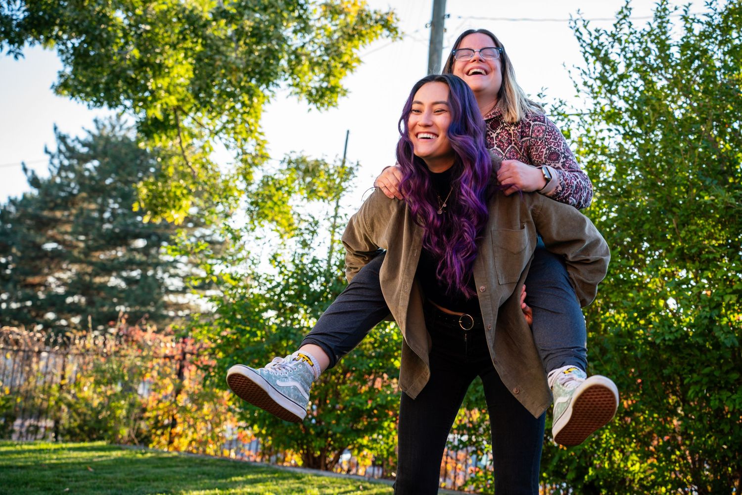 Two women laughing outdoors in autumn park, one giving the other a piggyback ride.