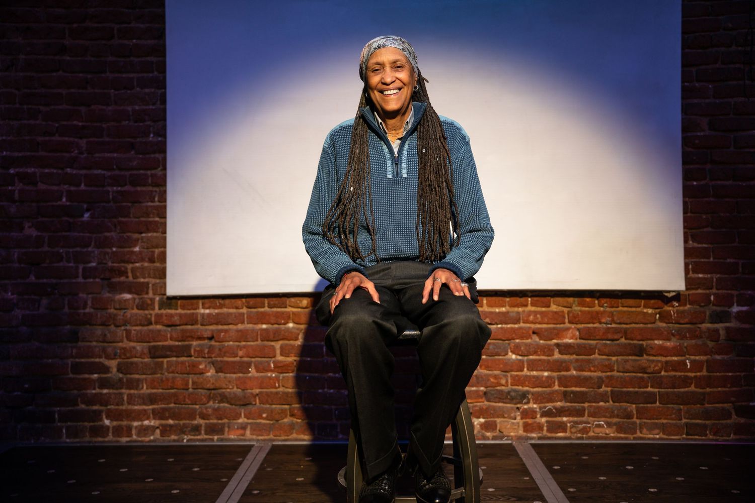 Older woman with long gray locs wearing a blue jacket, sitting and smiling in front of a brick wall.