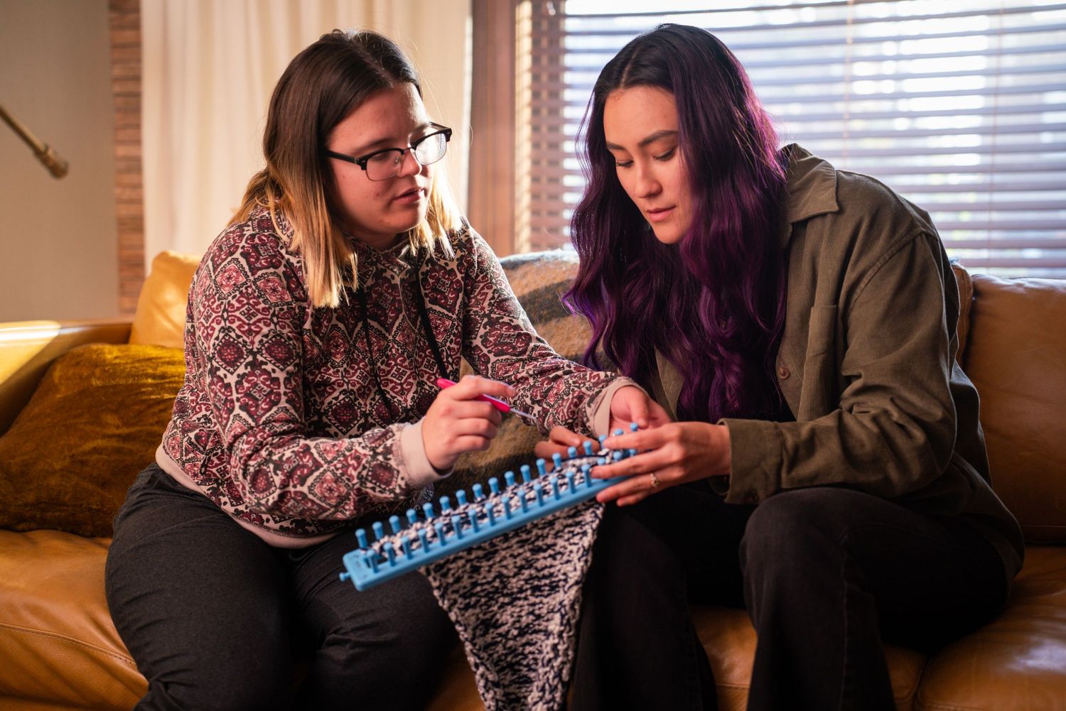 Two young women sit on a couch together, one teaching the other how to use a knitting loom.