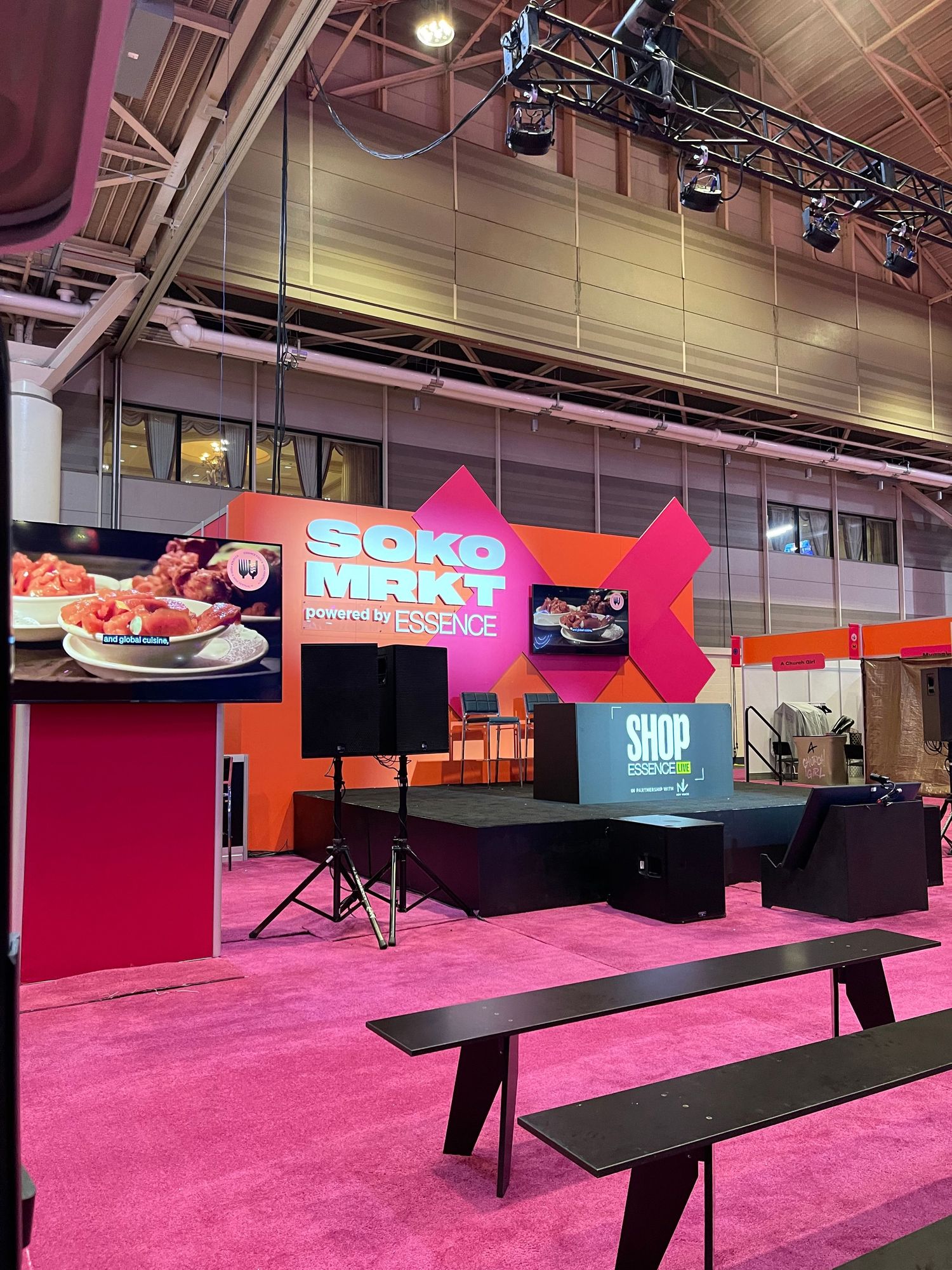 Soko Mrkt stage powered by Essence with a Shop Essence sign, black bench seating, and bright pink carpet inside a convention hall.
