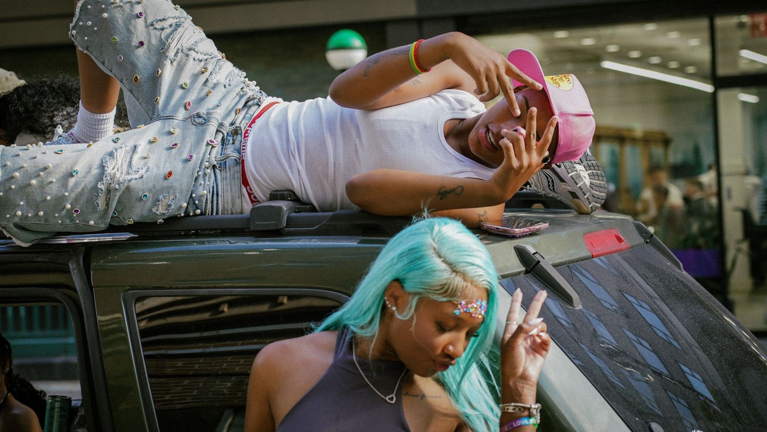 Two young women celebrate at Pride parade, one lying on car roof, one flashing peace sign.