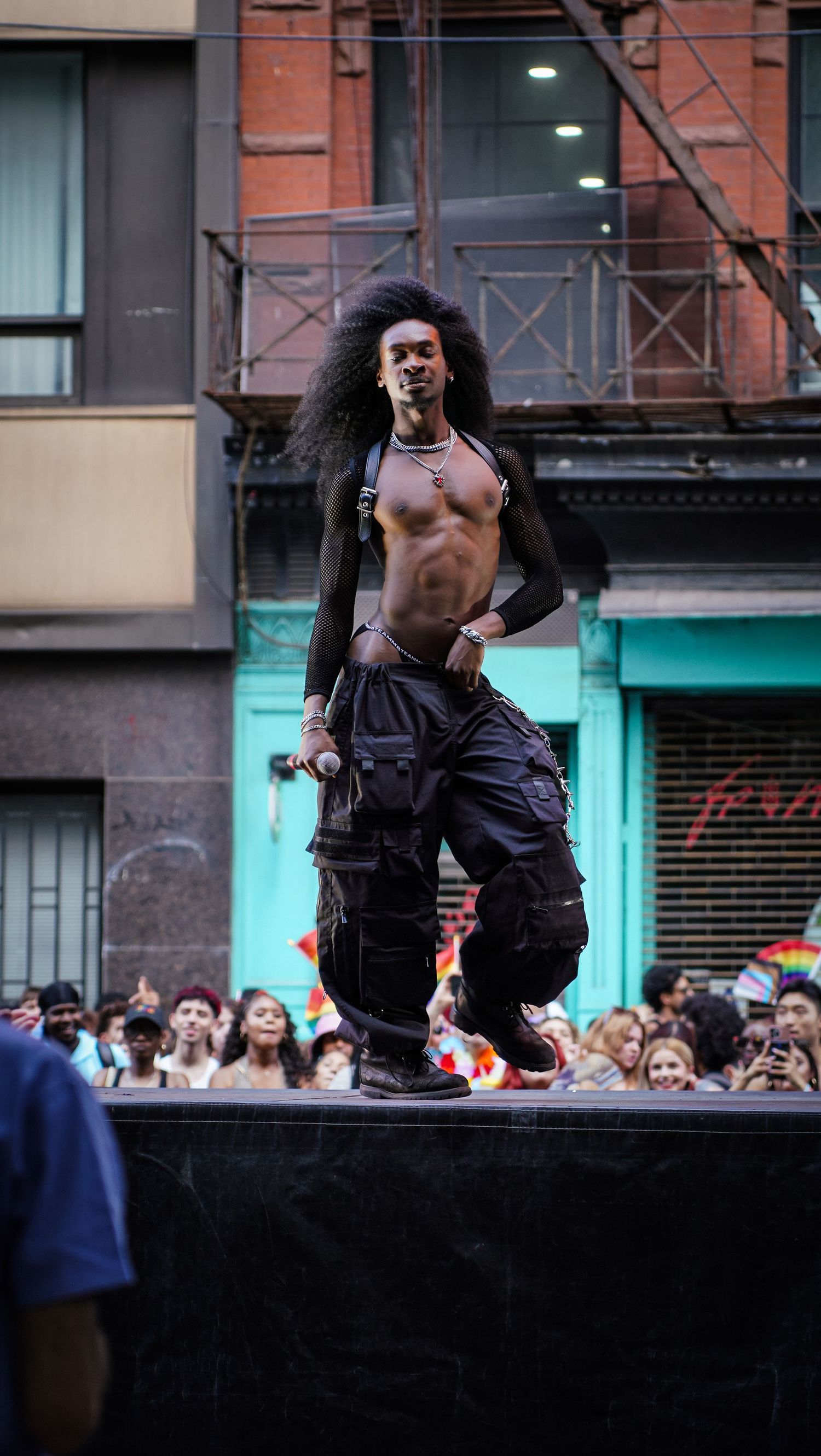 Shirtless Black performer with long curly hair poses confidently on stage at Pride celebration.