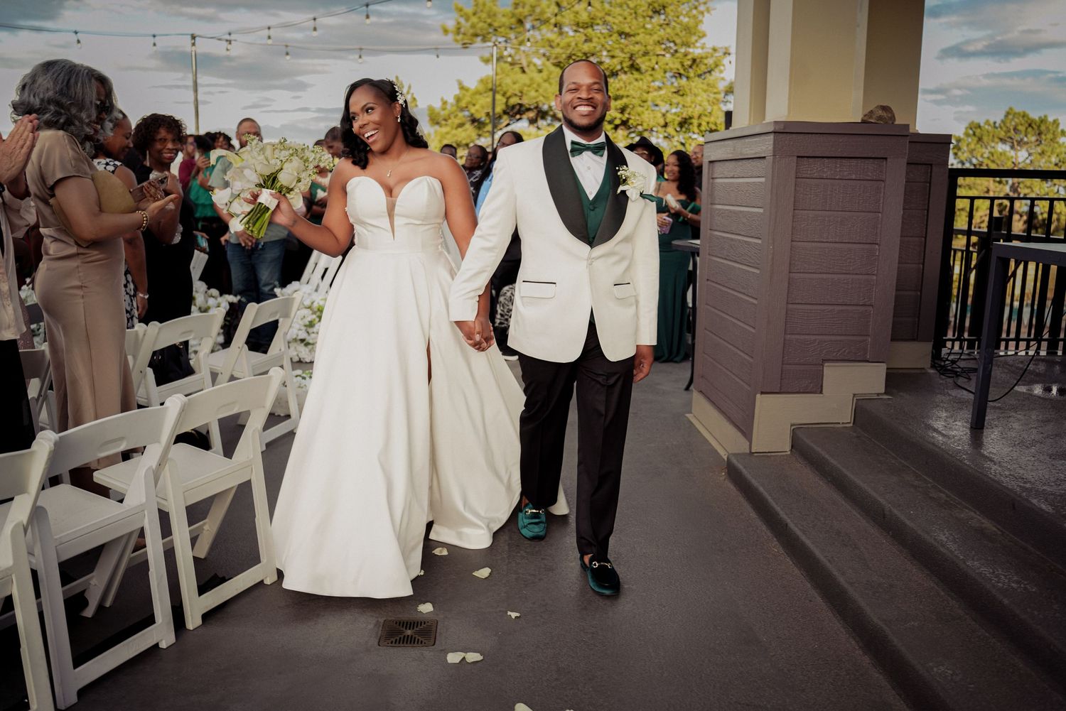 Newlywed couple walks down outdoor wedding aisle, bride in white gown, groom in white tuxedo, guests seated beside them.