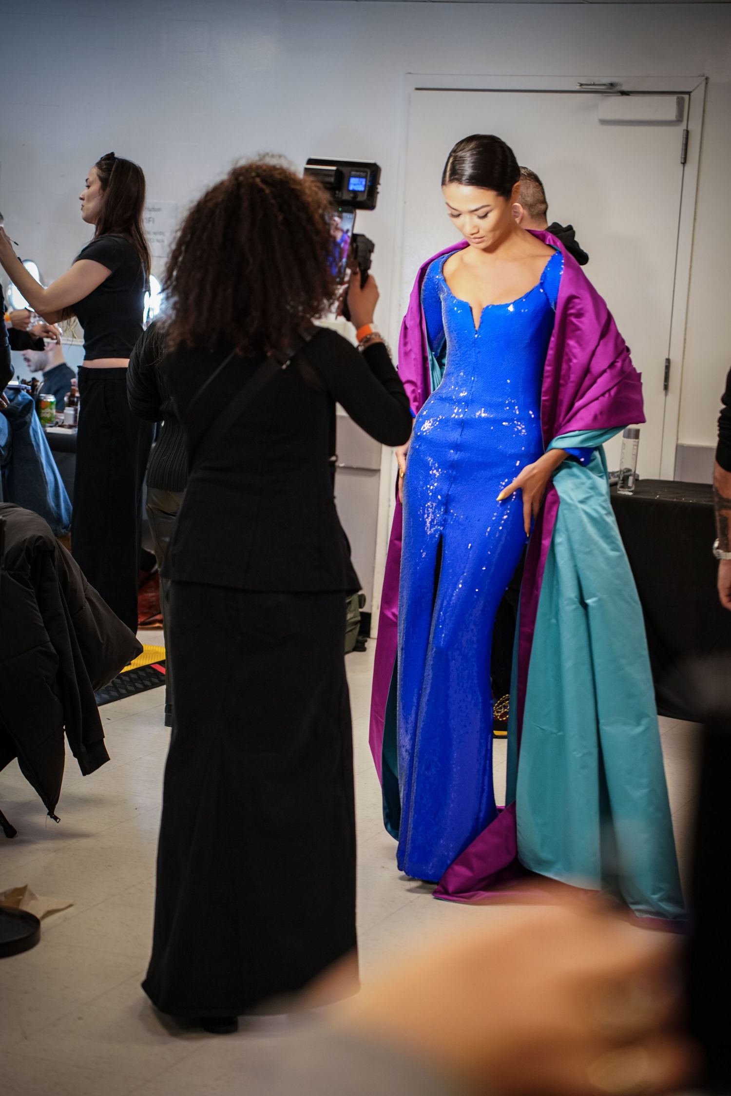 A model in a blue sequin gown with purple and teal cape being dressed backstage.