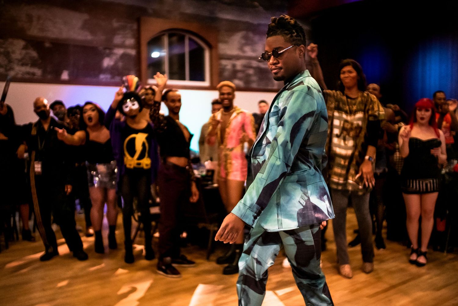 A man in a teal patterned suit dances confidently on a wooden floor surrounded by a crowd.