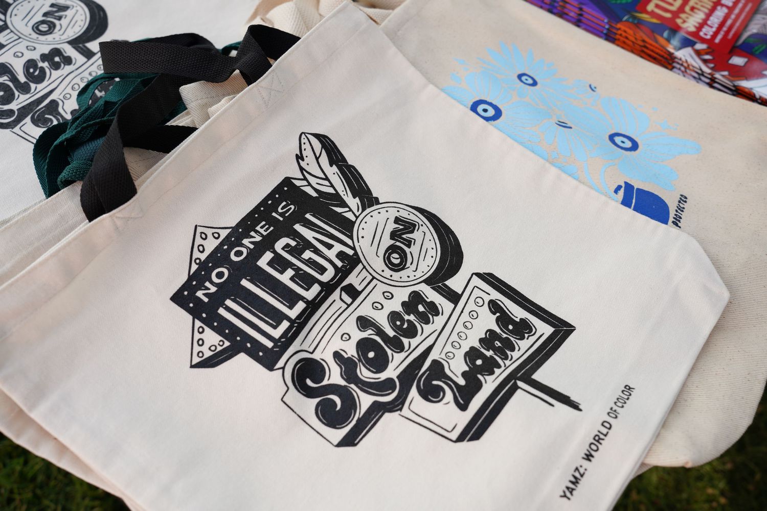 Canvas tote bag printed with No One is Illegal on Stolen Land at an outdoor Pride market.