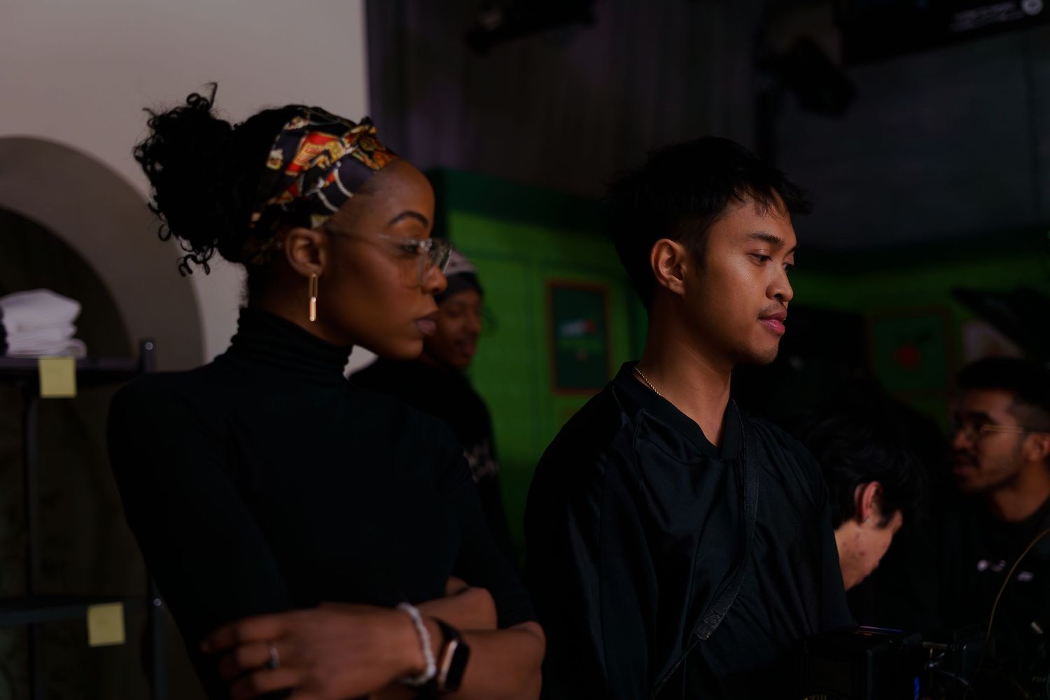 Two young creatives observe filming production in dimly lit venue with green ambient lighting in background.