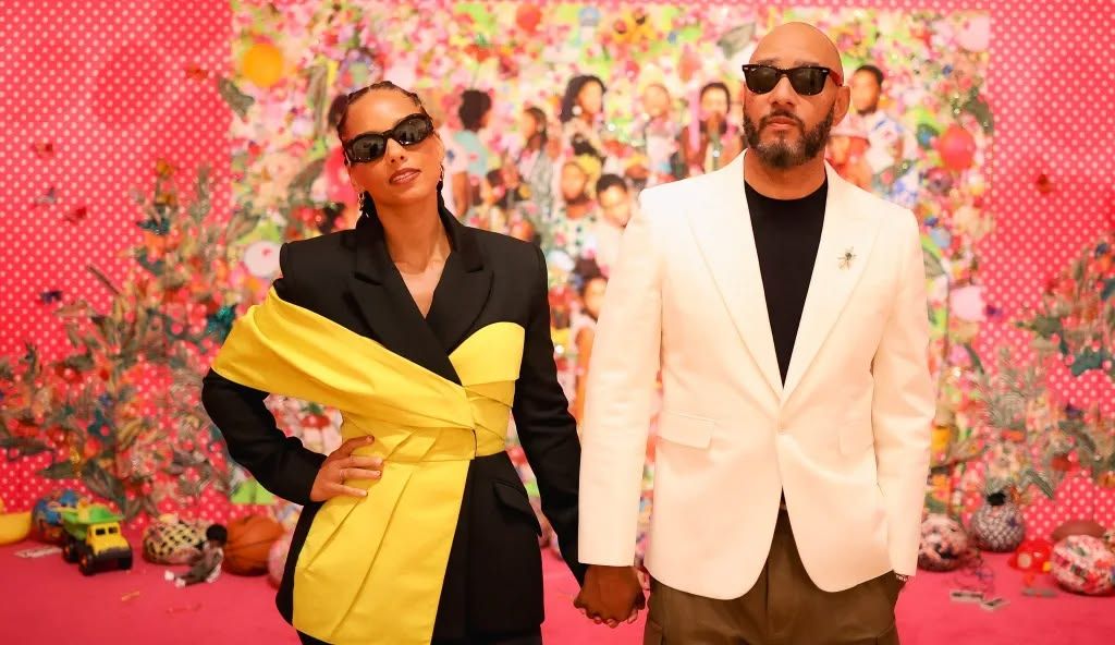 Two people in sunglasses hold hands in front of a colorful pink collage wall, one in a black blazer with yellow sash, one in a cream blazer.