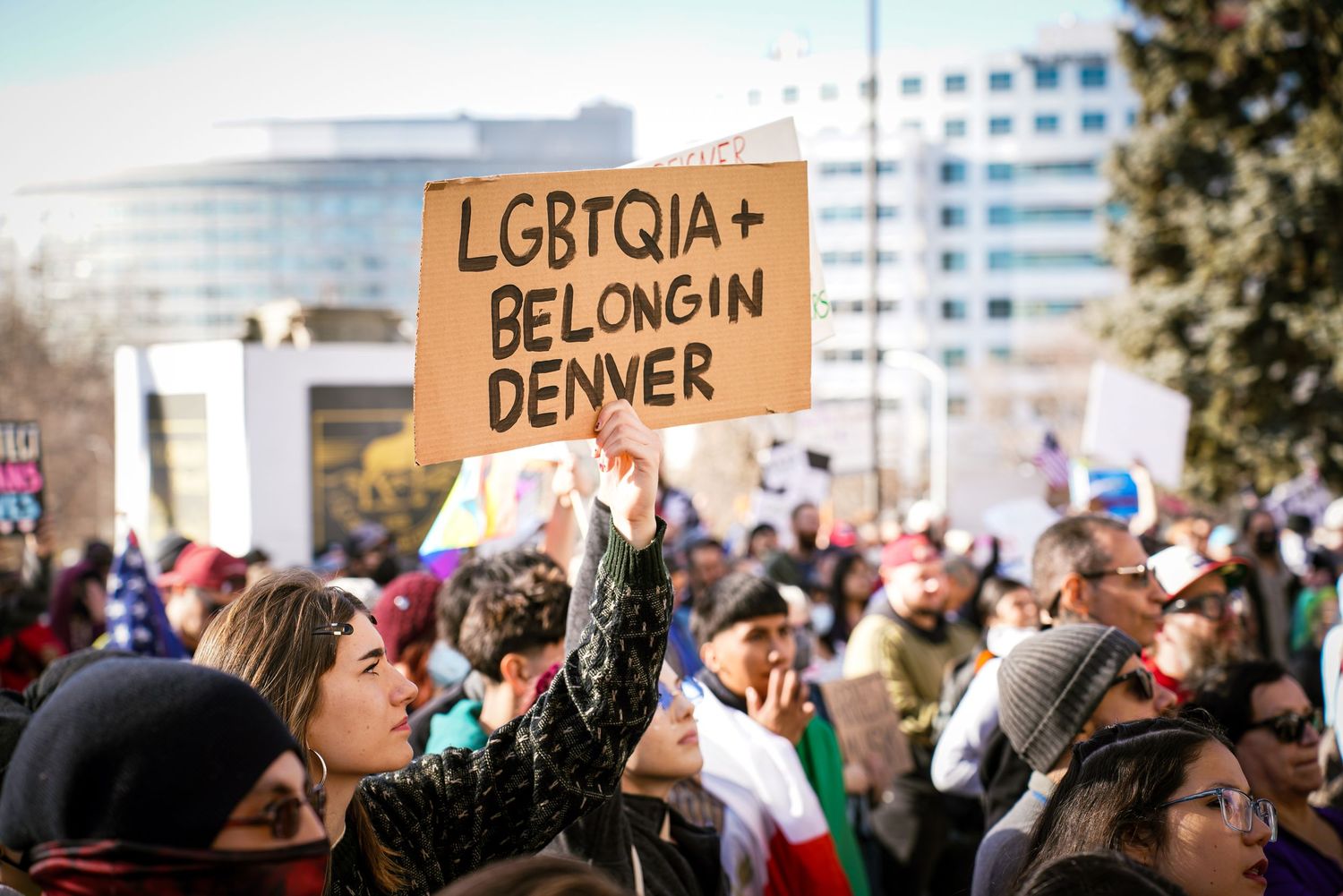 Crowd of protesters at Denver rally with person holding cardboard sign reading LGBTQIA+ Belong in Denver.